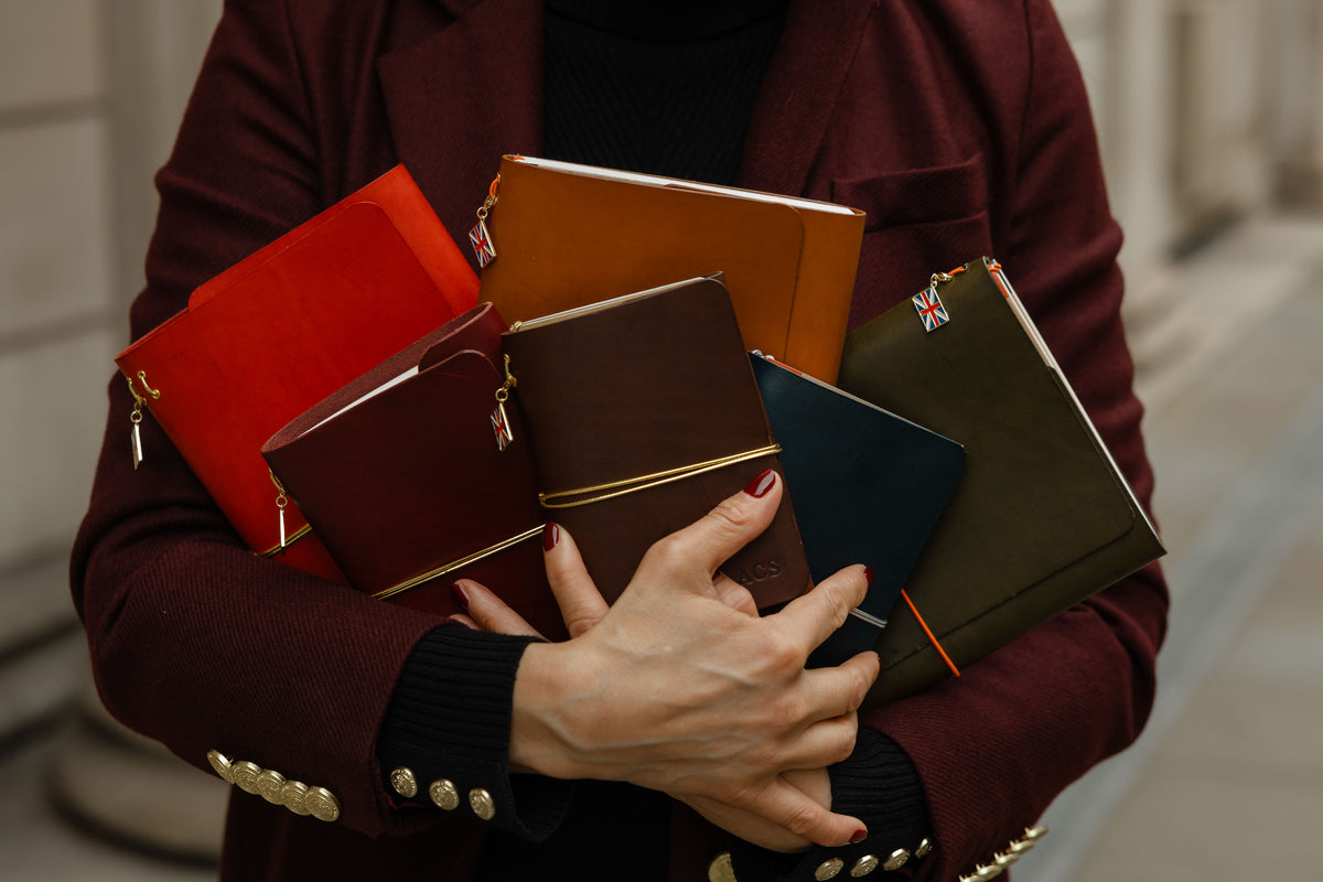 A person in a burgundy blazer holds a cluster of colorful leather notebooks and small zip pouches in their hands. The notebooks are in shades of red, orange, brown, navy, and olive green, with gold-tone zippers and snaps.